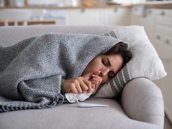 young woman lying under blanket on sofa coughing and sneezing using paper tissue to blow her nose unhealthy female caught cold or flu resting on couch suffering coronavirus