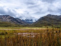a marshy lake reflecting the dramatic cloudy sky is surrounded by rolling hills and snow capped peaks on the pastoruri glacier trek in huaraz peru