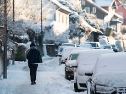 eine person geht auf einem verschneiten gehweg am strassenrand parken mit schnee bedeckte autos im tagesverlauf des sonntags herrscht auflockernde bewoelkung und oft sonne bei hoechstwerten von 7 grad