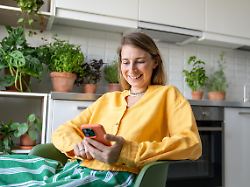 laughing woman engrossed in smartphone watching video news and social feed sit in kitchen surrounded by plants