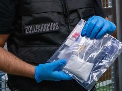 ein zollfahnder mit blauen plastikhandschuhen haelt eine plastikverpackung mit einem tuetchen mit weissem pulver kokain und rosanen pillen ecstasy in den haenden auf seiner weste steht zollfahndung