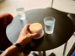hand reaches for a latte with a heart design on a small black table with two glasses of water