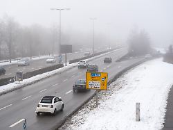 staedtischer verkehr auf einer breiten ausfallstrasse im winter autos fahren bei nebel und kaelte in richtung innenstadt waehrend schnee und glaette die aufmerksamkeit der verkehrsteilnehmer erhoehen