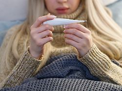 starke junge frau mit fieber liegt im bett unter der decke ueberprueft die temperatur und schaut auf thermometer selektive fokus