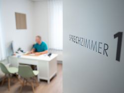 ein hausarzt sitzt in einem sprechzimmer seiner hausarztpraxis an einem schreibtisch und arbeitet am computer