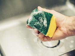 mann mit schwamm und reinigungsmittel reinigt waschbecken unter fliessendem wasser