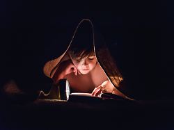 young boy reading a book under a blanket using a flashlight