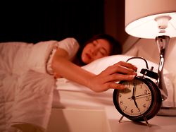 sleepy young woman in bed with eyes closed extending hand to alarm clock in the morning