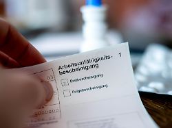 eine person haelt eine ausgefuellte arbeitsunfaehigkeitsbescheinigung in der hand im hintergrund ist unscharf ein tisch mit medikamenten zu sehen