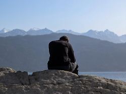 a man sits alone on the rocky shoreline of lake walchensee in urfeld bavaria germany on march 7 2026 snow covered peaks of the bavarian alps rise in the background above the alpine lake landscape