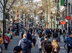 diese aufnahme zeigt das belebte treiben von zahlreichen passanten auf der sonnendurchfluteten einkaufsmeile schildergasse in koeln gesaeumt von markanten ladenfronten und baeumen