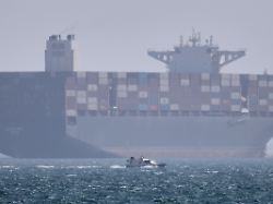 ein containerschiff liegt vor anker waehrend im vordergrund ein kleines motorboot in der strasse von hormus vor vorbeifaehrt