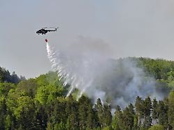 ein hubschrauber fliegt mit einem loeschwasser behaelter ueber ein waldgebiet in dem es im nationalpark boehmische schweiz brennt