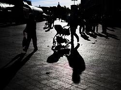 ein mann schiebt im gegenlicht der sonne einen kinderwagen durch die innenstadt und wirft dabei einen langen schatten auf den boden