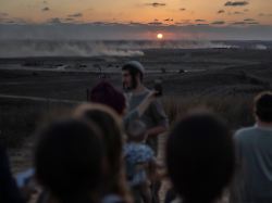 rechtsgerichtete israelische siedler versammeln sich an der grenze zum gazastreifen waehrend im hintergrund rauchschwaden von vorrueckenden israelischen panzern zu sehen sind