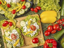 zwei brotscheiben dekorativ belegt mit rohkost und frischkaese auf holzbrett umgeben von tomaten paprika und salat