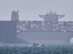 archiv 02 05 2026 iran bandar abbas ein containerschiff liegt vor anker waehrend im vordergrund ein kleines motorboot in der strasse von hormus vor vorbeifaehrt