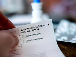 eine person haelt eine ausgefuellte arbeitsunfaehigkeitsbescheinigung in der hand im hintergrund ist unscharf ein tisch mit medikamenten zu sehen
