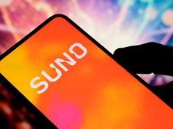 in this photo illustration the suno logo is displayed on a cell phone held in one hand by a man with an illustration of a concert atmosphere in the background in creteil france on november 27 2025