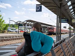 symbolbild junge frau mit rucksack und nackenkissen wartet am bahnhof 29 06 2026 eine reisende sitzt mit gepaeck und nackenkissen im bahnhofsbereich und wartet auf ihre zugverbindung