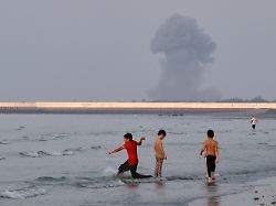 three boys play in the shallow waters of the strait of hormuz as a plume of smoke rises from an explosion in the background off bandar abbas iran monday july 13 2026
